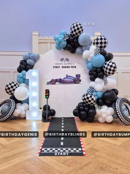 A "First Grand Prix" theme with a clean and stylish design. The setup includes a starting line floor runner, a traffic light prop, and a balloon arch in black, white, and blue.