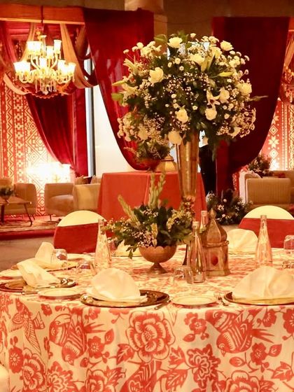 An elegant table setting for the "Noor-e-Kashmir" event, featuring custom printed tablecloths and tall floral centerpieces, all within a space draped in rich red fabrics.