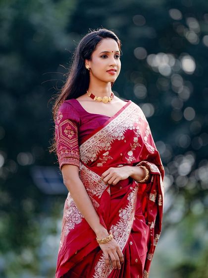 A beautiful outdoor portrait of a woman in a rich, red embroidered saree. The natural background complements the traditional attire.