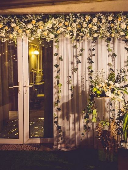 An elegant floral backdrop designed for an evening event. A cascade of white flowers and trailing ivy over sheer curtains creates a soft, romantic photo wall, complemented by modern floral stands.