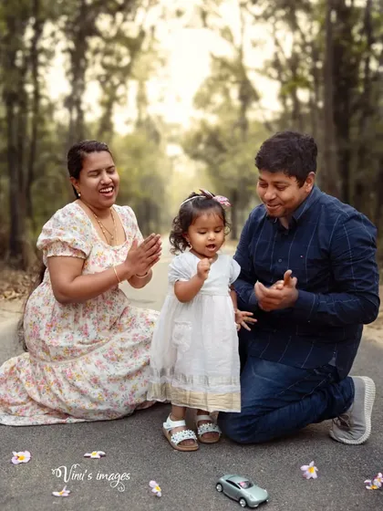 Pure family joy captured outdoors. This candid moment shows parents clapping and smiling with their toddler on a tree-lined road, creating a memory full of laughter and love.