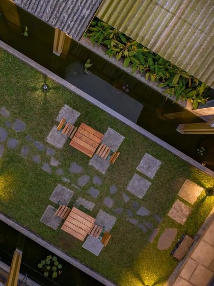 An aerial view of the outdoor seating area, showing the geometric pattern of stone pavers on the lawn. This space is flanked by water bodies and sheltered by the overhanging corrugated roofs, creating a perfect spot for enjoying tea.