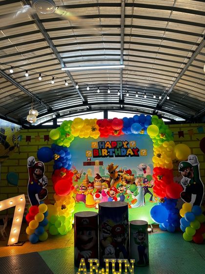 This Super Mario theme brings the iconic video game to life with a full backdrop of the Mushroom Kingdom. The setup is framed by a rainbow balloon arch and features character cutouts of Mario and Luigi, along with a light-up marquee number '7'.