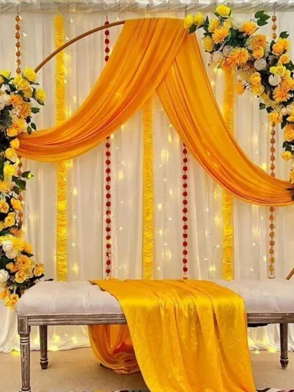 An elegant Haldi seating arrangement with a white bench and a circular arch. The arch is decorated with yellow flowers and draped with yellow fabric, set against a backdrop of white curtains and fairy lights.