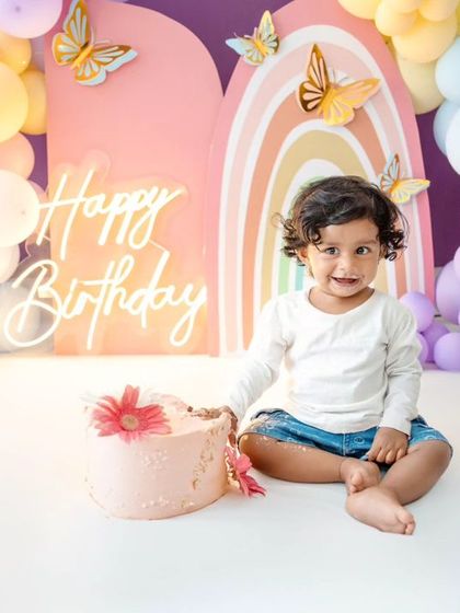 A sweet portrait of the birthday girl beside her cake, ready to celebrate in front of the colorful butterfly backdrop.