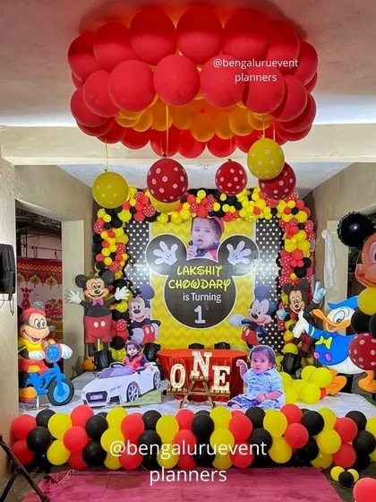 This is a complete Mickey Mouse themed first birthday party I created. The setup includes a personalized backdrop with the child's photo, multiple character cutouts, and coordinated red, yellow, and black balloon decorations for a fun and immersive experience.