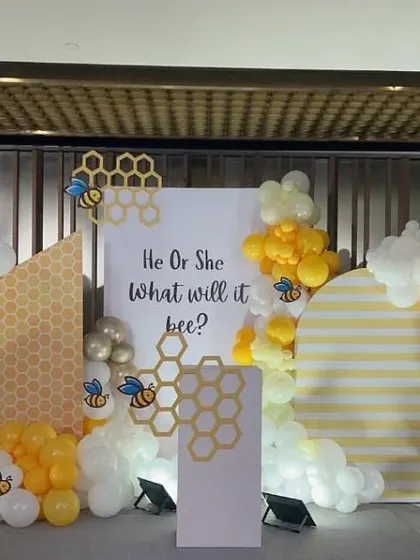 A unique and adorable 'He or She, What will it bee?' gender reveal theme. The backdrop features a honeycomb pattern with yellow, white, and gold balloons and cute bee accents for a playful touch.