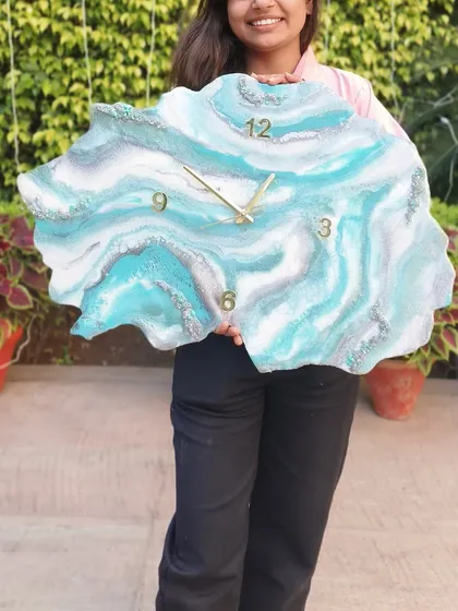 A large, free-form geode clock held by a happy student. The flowing patterns of turquoise, white, and silver resin mimic the natural layers of a stone agate.