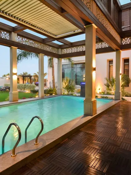 A sun kissed pool area under a retractable louvre roof. The design combines wooden decking, intricate trellis work, and classical pillars to create a luxurious and harmonious outdoor space.