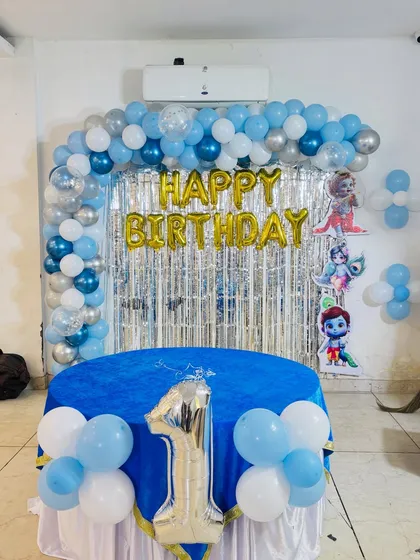 A first birthday party setup with a cartoon character theme. The decor includes a blue and silver balloon arch, a silver tinsel backdrop, and a decorated table for the cake.