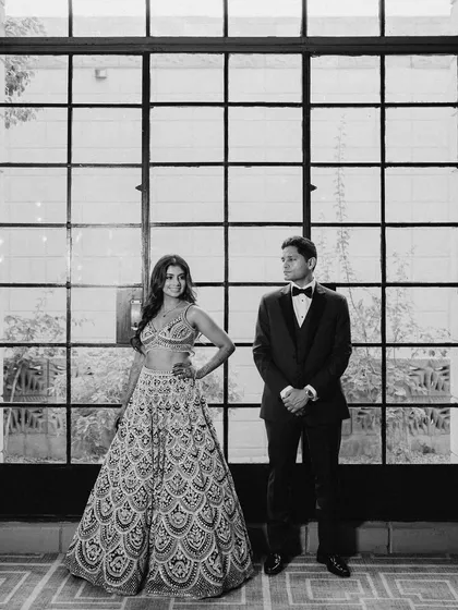 A playful and elegant black and white portrait. The couple stands against a large window, their contrasting poses creating a dynamic and stylish image.