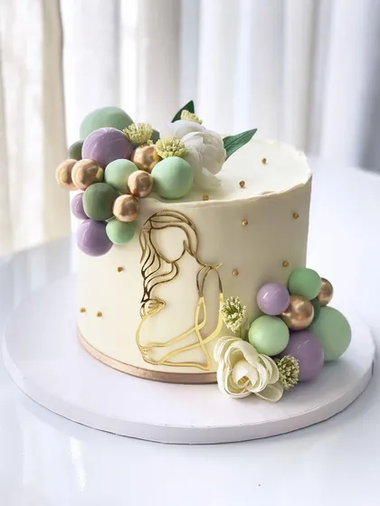 A beautiful baby shower cake with a modern, artistic silhouette of a mother-to-be, accented with a cascade of green, purple, and gold chocolate spheres.