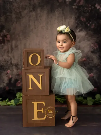 A proud one-year-old stands next to her birthday blocks, showing off her new skill of standing.