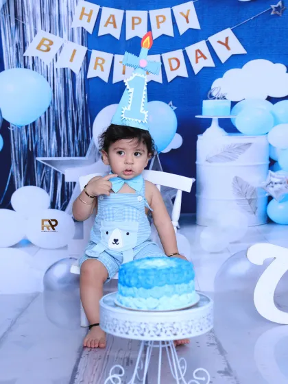 A sweet moment during a cake smash session, where the birthday boy sits calmly in front of his cake. The blue and white setup with a "Happy Birthday" banner creates a classic party atmosphere.