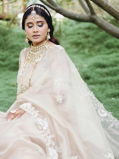 A serene portrait of the bride, Aishwarya, seated in a garden. The soft light and her elegant, neutral-toned saree create a peaceful and ethereal image.
