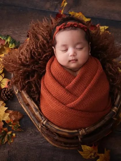 Cozy autumn vibes for a new arrival. This newborn is wrapped snugly in a rust-colored swaddle, nestled in a basket with autumn leaves. The warm colors and textures create a beautiful, seasonal portrait.