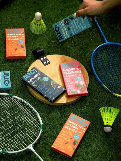 A playful, sporty flatlay for Alt Co. chocolates. The badminton rackets and shuttlecocks create a fun, active context, suggesting these are a great post-activity treat.