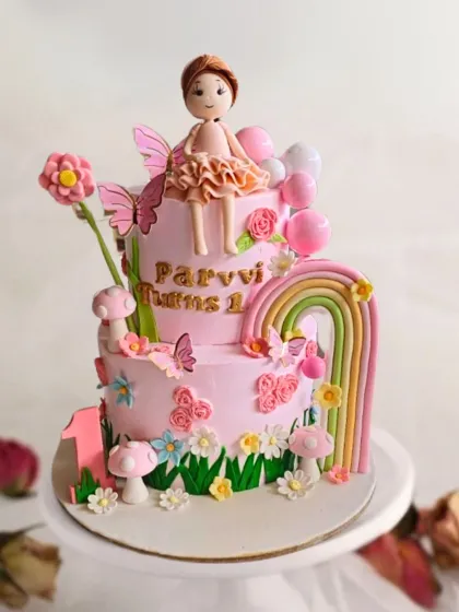 A two-tier fairytale cake featuring a delicate fairy, a rainbow, and a garden of flowers and butterflies for a magical first birthday.