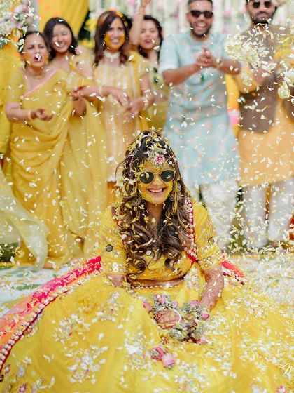 The bride, looking cool and happy in her sunglasses, gets showered with petals during her vibrant Haldi ceremony.