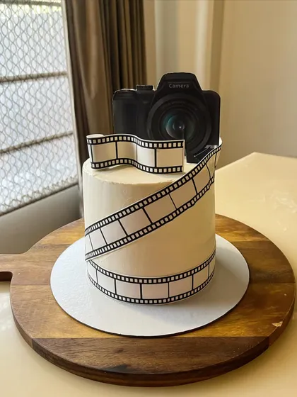 The perfect cake for a photography lover. This design features an edible film strip wrapped around a buttercream cake, topped with a detailed camera figure.