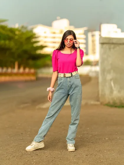 A full-length fashion portrait, with the model adjusting her sunglasses, showcasing a cool and casual pink and denim outfit.