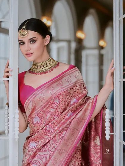 A model framed by a white doorway, wearing a beautiful pink Banarasi silk saree. The composition feels intimate, as if peeking into a private moment of elegance.