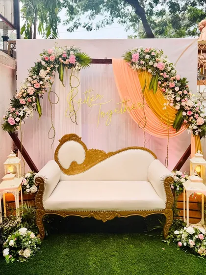 A simple yet elegant photo booth for an intimate function. The design uses a floral arch, soft drapes, and a "Better Together" neon sign, with a classic diwan for seating.