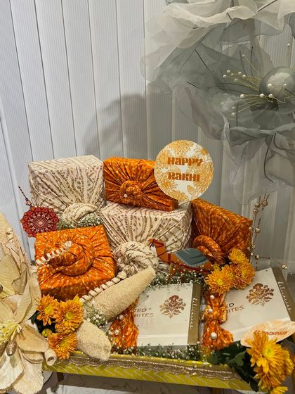 A close-up of the orange-themed traditional Rakhi platter. The fabric-wrapped gifts are adorned with small flowers and tags.