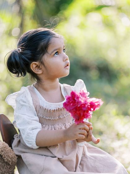 A little girl holding a pink flower, looking up thoughtfully. A beautiful, candid portrait of a child lost in her own world.