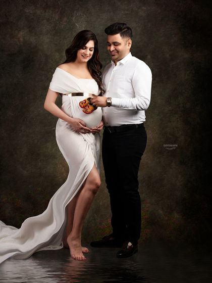 A sweet and classic couple shot. The parents-to-be hold tiny baby shoes over the bump, symbolizing their excitement. The white gown and dark background create a timeless look.