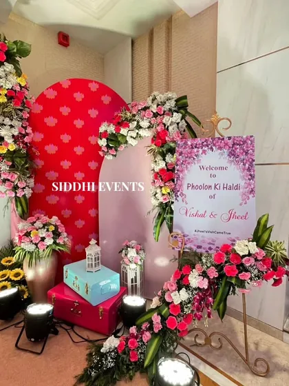 The welcome corner for the 'Phoolon ki Haldi', featuring a personalized sign, colorful trunks, and an abundance of pink, white, and yellow flowers.