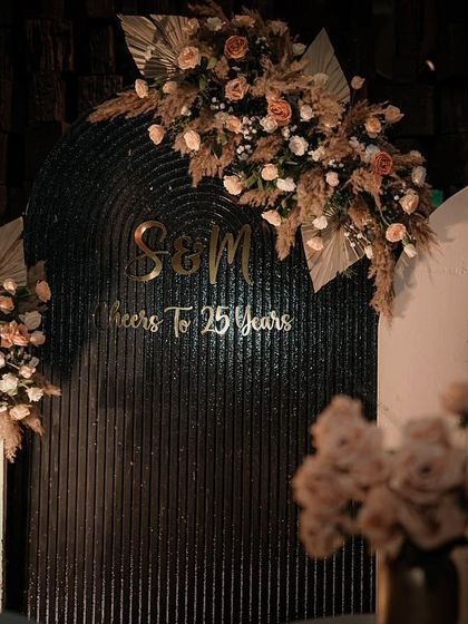 A close-up of the personalized decor for a 25th-anniversary celebration. The couple's initials and a "Cheers to 25 Years" message are set against a modern, textured backdrop with elegant floral sprays.