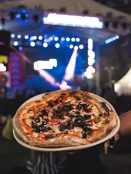 Holding up a freshly made pizza against the backdrop of a live concert stage. Our catering service brings the best food to the best events.