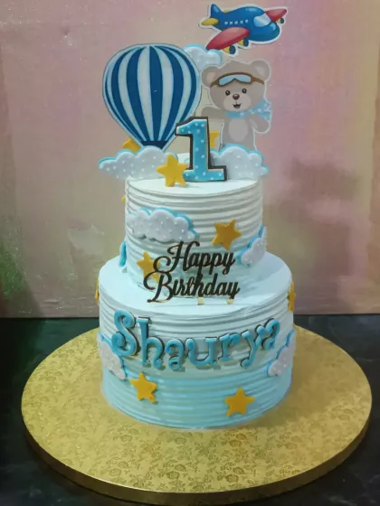 A cheerful two-tiered cake for a first birthday, with a hot air balloon and airplane theme. The cute teddy bear pilot and fluffy clouds make it perfect for a little adventurer.