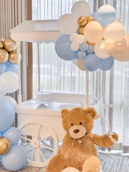 We use charming props to enhance our themes. Here, a large, cuddly teddy bear sits next to a white cart, which is perfect for holding gifts or a cake, all tied together with a blue, white, and gold balloon arrangement.