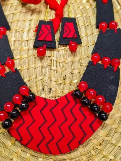 This bib-style necklace uses a bold chevron pattern on red fabric, contrasted with black panels and beads. The geometric design and the striking red and black color combination make it a powerful statement piece. It's designed to be surprisingly lightweight and comfortable.