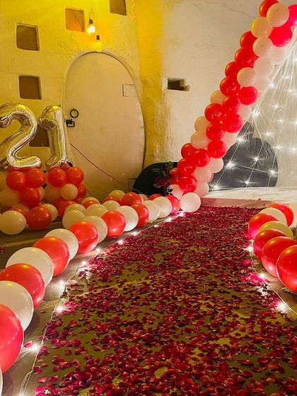 A unique 21st birthday setup in a rustic-style room. The decor includes a path of rose petals, a fairy light tent, and red and white balloons.