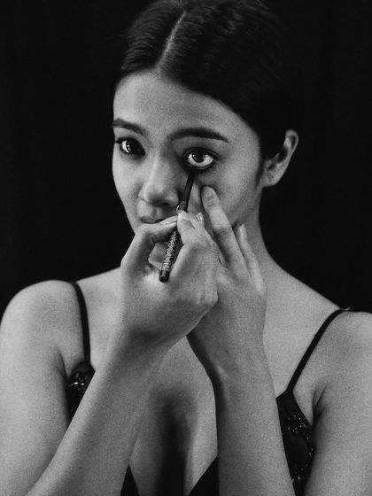 An intense moment of focus, captured in high-contrast black and white. The model applies her eyeliner while looking directly at her reflection, and the dark background isolates her, making the moment feel personal and dramatic.