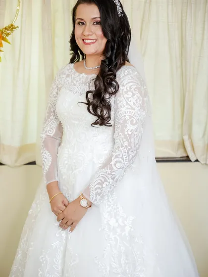 A happy and radiant bride in her long-sleeved lace gown. Her hair is styled in soft curls, and her makeup is natural and glowing.