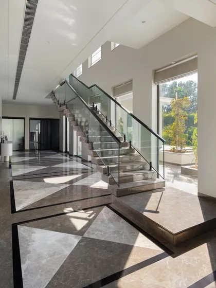 Sunlight streams into this hallway, creating a beautiful play of shadows across the geometric inlaid marble floor. The modern glass-railed staircase appears to float, enhancing the sense of light and space.