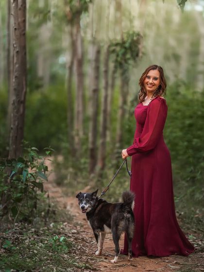 A woman in a flowing burgundy dress walks her dog on a forest trail. This full-length outdoor portrait captures a simple, elegant moment between a pet parent and her companion.