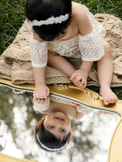 A baby girl looks at her reflection in a vintage mirror placed in the grass. A creative and beautiful concept for a sitter session.