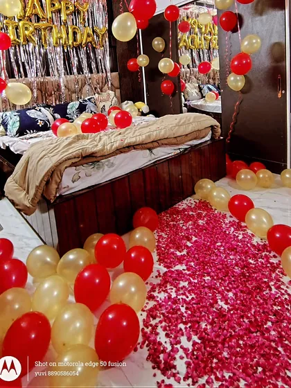 This photo shows the romantic details of a birthday room setup. A path of red and white rose petals leads to the bed, which is decorated with balloons and a 'Happy Birthday' backdrop.