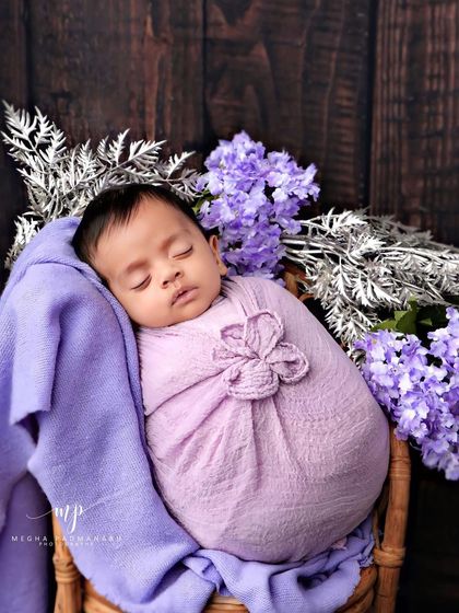 A baby in a purple flower-style wrap, posed on a rattan chair against a dark wood background.