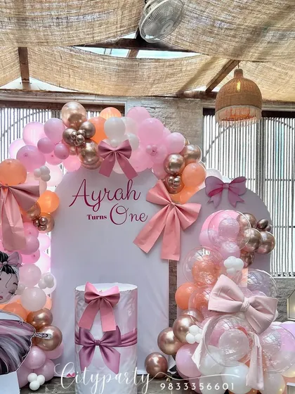 This elegant bow-themed first birthday decoration uses a palette of pink, peach, and rose gold. The setup is detailed with large fabric bows, a custom backdrop, a light-up marquee number, and a character standee.