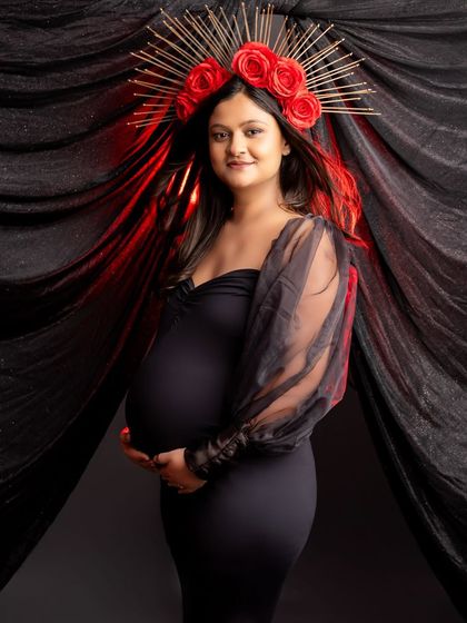 A powerful solo portrait with a touch of red. The mom-to-be wears a striking red floral crown that contrasts beautifully with her black dress and the dark, draped background.