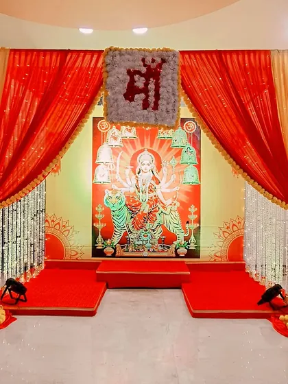 A traditional mandap setup for Durga Puja. The design features a classic red and gold color scheme, a beautiful backdrop of Maa Durga, and floral "Maa" lettering, creating a sacred space for worship.