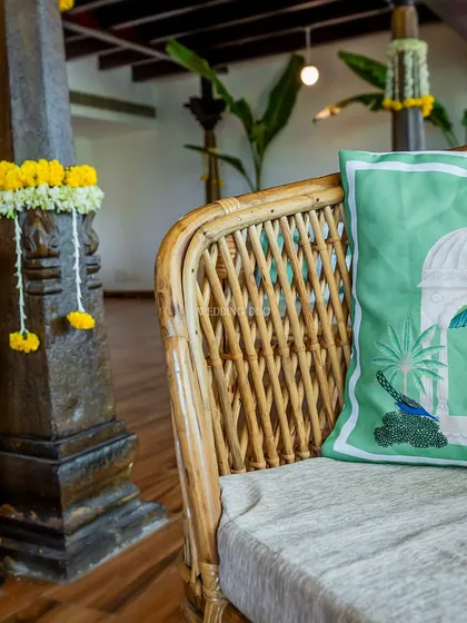 A detail of our Haldi decor, where a traditional stone pillar is adorned with marigold garlands, next to a cane sofa with a custom peacock-print cushion.