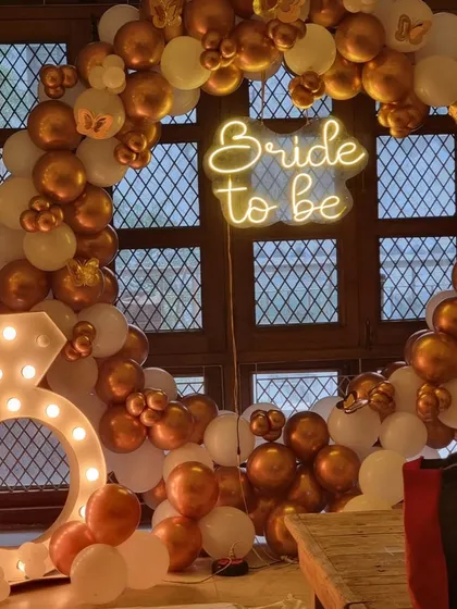 Celebrate the bride-to-be in style. This chic bachelorette party decoration features a "Bride to be" neon sign on a gold and white balloon arch, complete with a large light-up diamond ring prop for fun photos.
