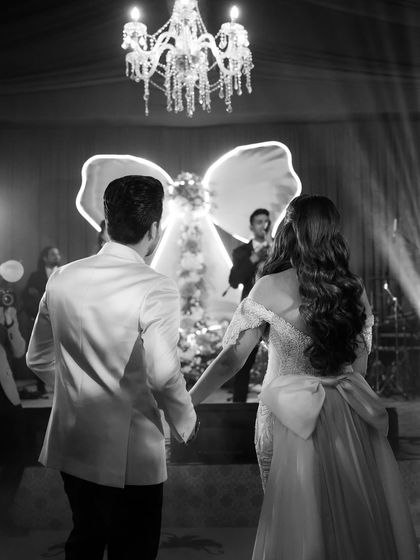 A beautiful black and white shot of the couple walking towards the stage, with the grand chandelier and statement bow creating a dramatic and romantic silhouette.